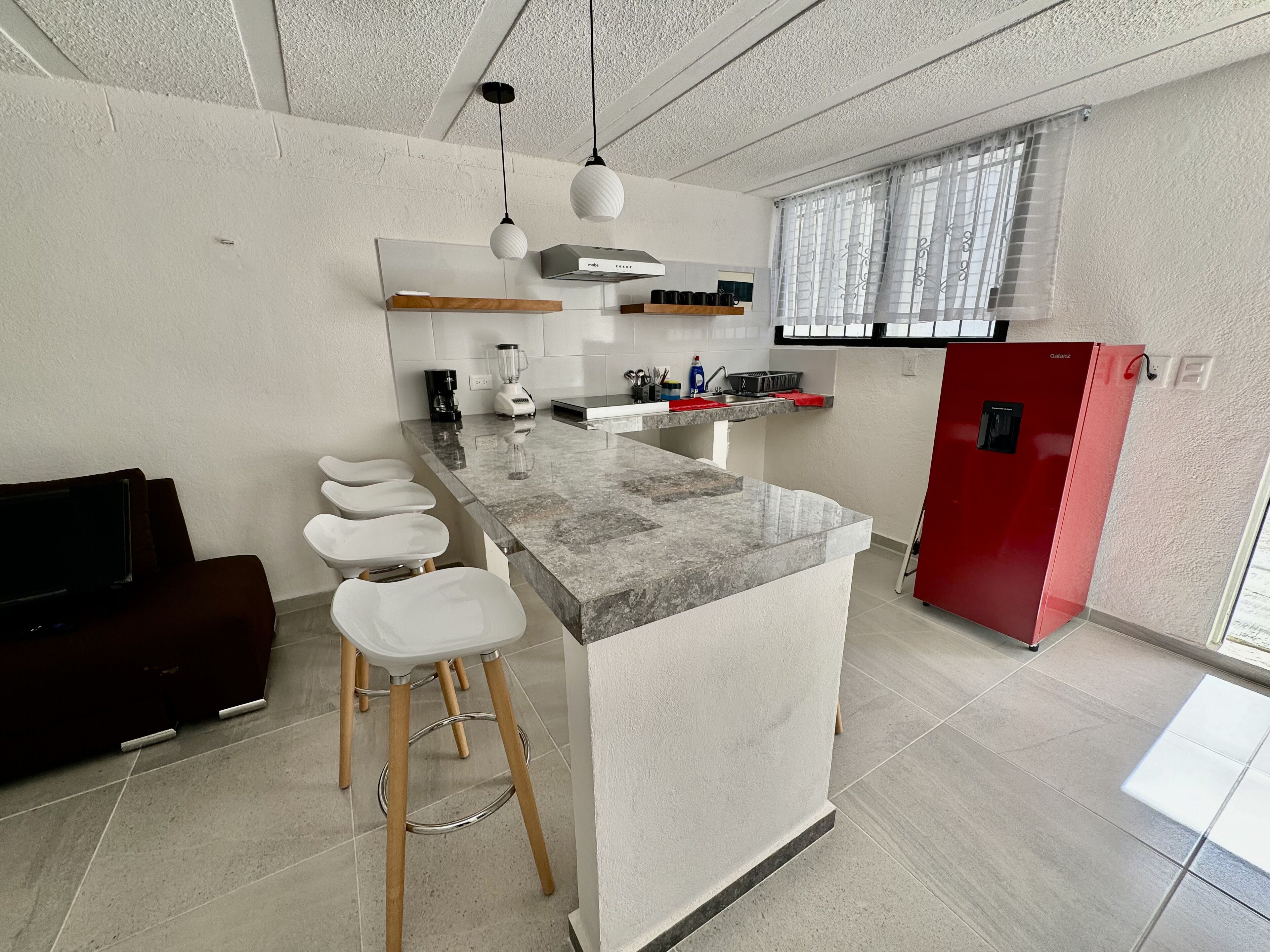 A modern kitchen with a marble countertop, wooden stools, a red refrigerator, and a built-in oven.