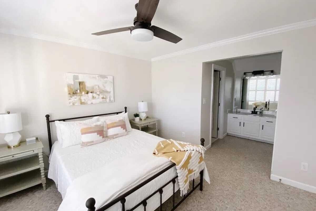 A bedroom with a bed, bedside tables, a ceiling fan, a painting on the wall, and a view into the bathroom.