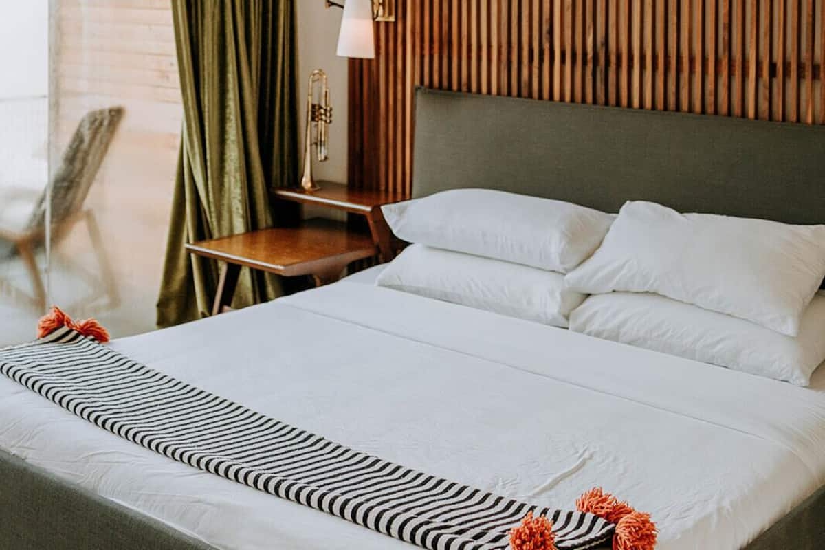 A modern hotel room with a bed, a wooden slat headboard, a hanging light fixture, and a view of the outdoors.