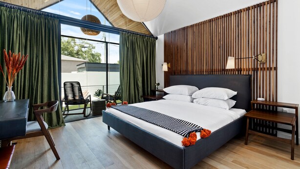 Ein modernes Schlafzimmer mit einem großen Bett, einem Schreibtisch und einem Stuhl. Ein Fenster mit Vorhängen, eine Wand aus Holzlamellen und eine Pendelleuchte sind ebenfalls vorhanden.