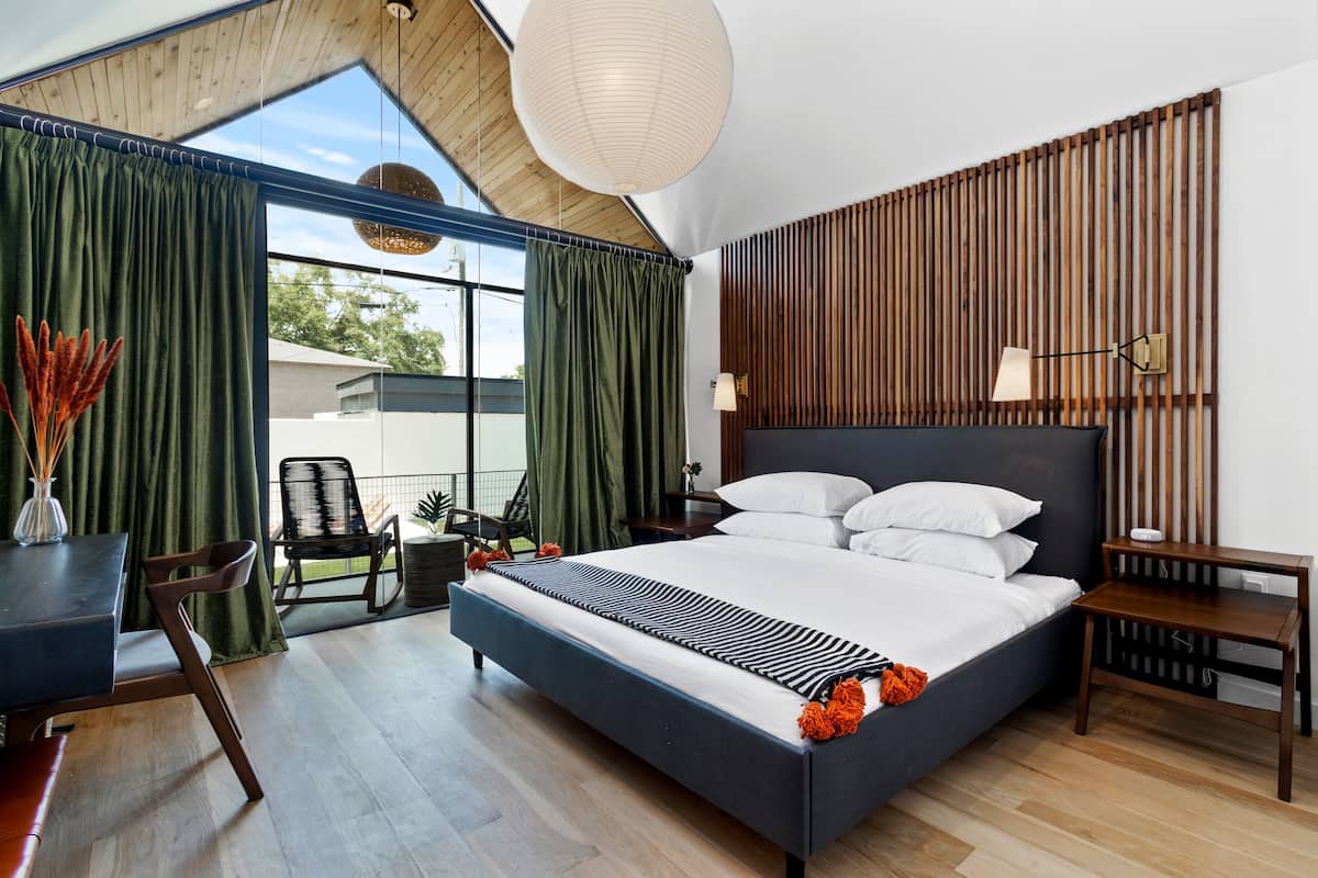 A modern bedroom with a large bed, a desk, and a chair. There is a window with curtains, a wooden slat wall, and a hanging light fixture.