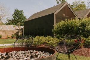 A patio area with two black wire chairs, a fire pit with rocks, and a wooden building with a sloped roof in the background.