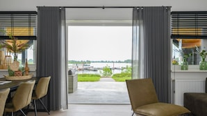 A modern living room with a view of a river and boats, featuring a dining area with chairs and a table, and a large window with curtains. - Nice home in Kerkdriel with WiFi (Kerkdriel)