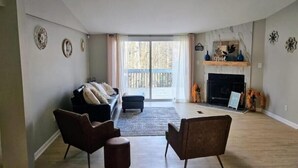 Ein geräumiges Wohnzimmer mit Kamin, einer Couch, zwei Sesseln und einem großen Fenster mit Blick auf Bäume.