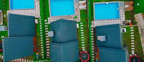 An aerial view of a residential area with three swimming pools, each surrounded by lounge chairs and umbrellas. There are three large buildings with flat roofs, and a circular trampoline is visible near the right building.