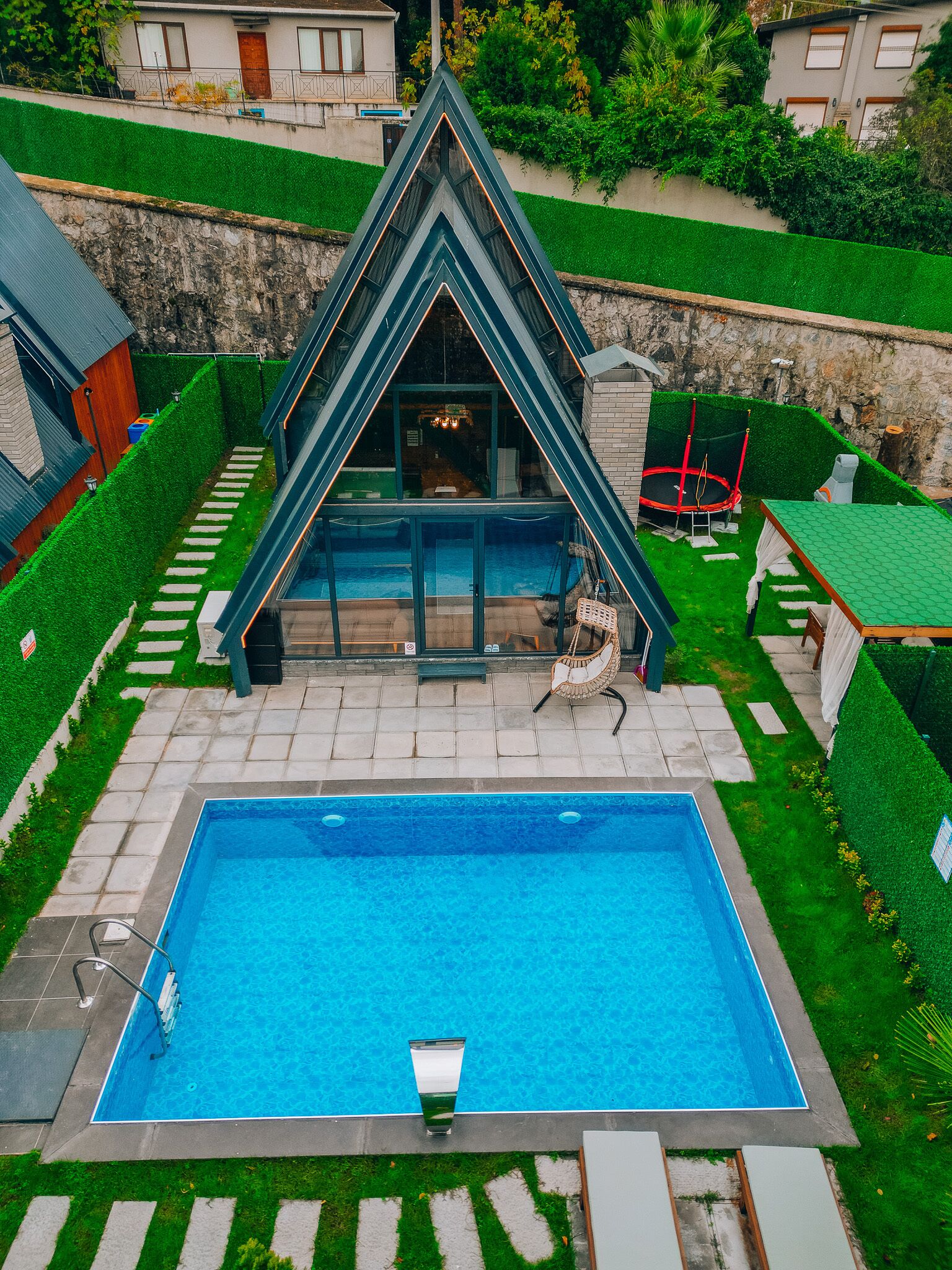 A modern swimming pool with a geometric glass enclosure, surrounded by a well-manicured lawn and a stone wall.