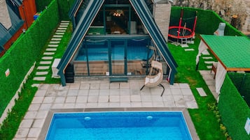 Una piscina moderna con cerramiento de vidrio geométrico, rodeada de césped cuidado y un muro de piedra.