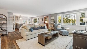 Un salon spacieux avec un grand canapé, une table basse et un fauteuil à motifs. Il y a également un coin cuisine avec des meubles blancs et une table à manger.