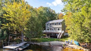 Une maison à deux étages avec un quai s’avançant dans un lac paisible, entourée d’une forêt dense.