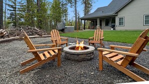 En eldstad med fyra trästolar i Adirondack-stil, placerad på ett område med grus, omgiven av träd och ett hus i bakgrunden.