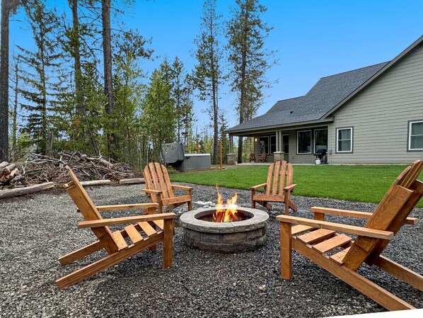 A fire pit with four wooden Adirondack chairs in a gravel area, surrounded by trees and a house in the background.