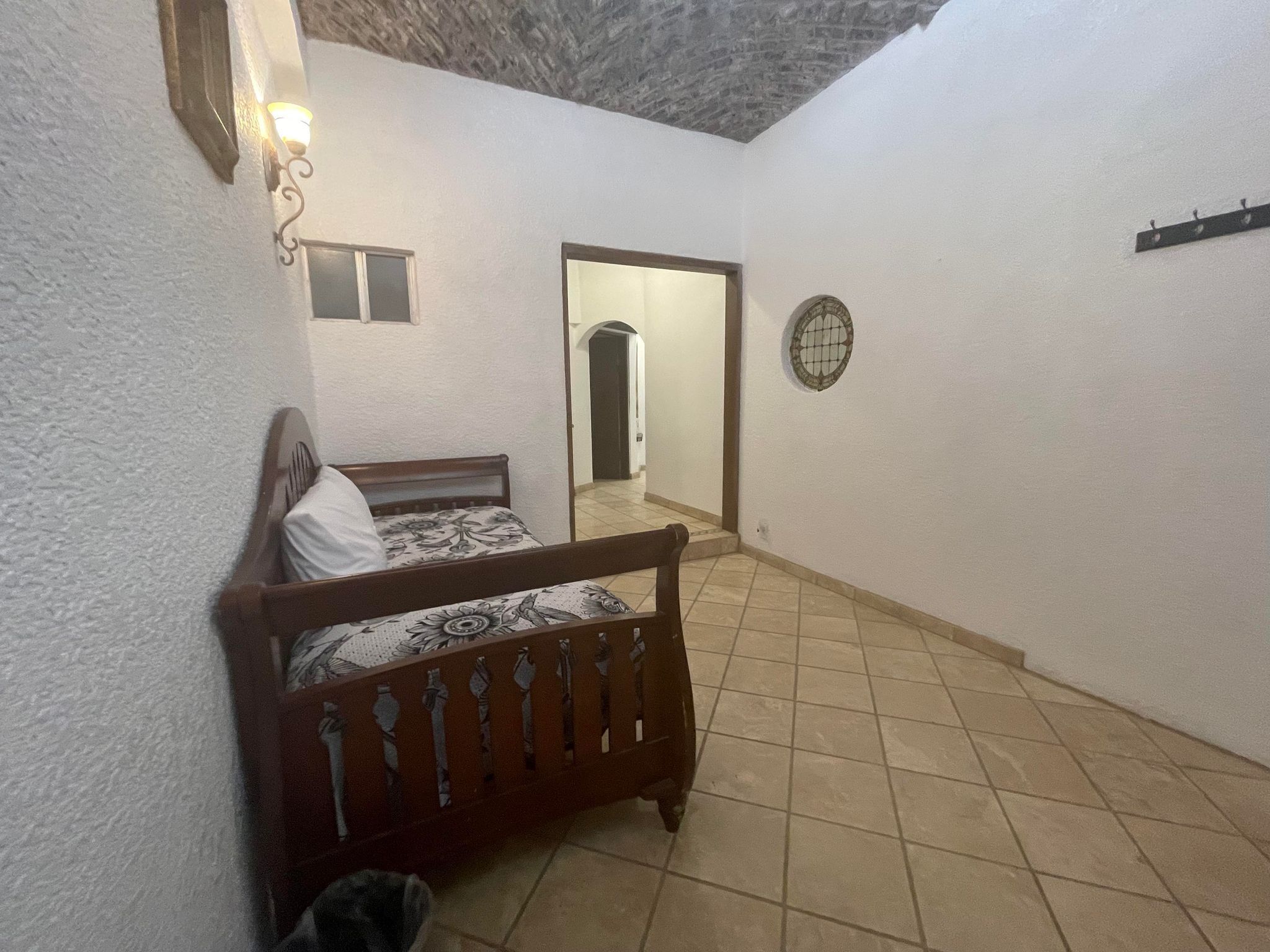 A bedroom with a wooden bed, a small mirror, a wall-mounted light fixture, and a tiled floor.