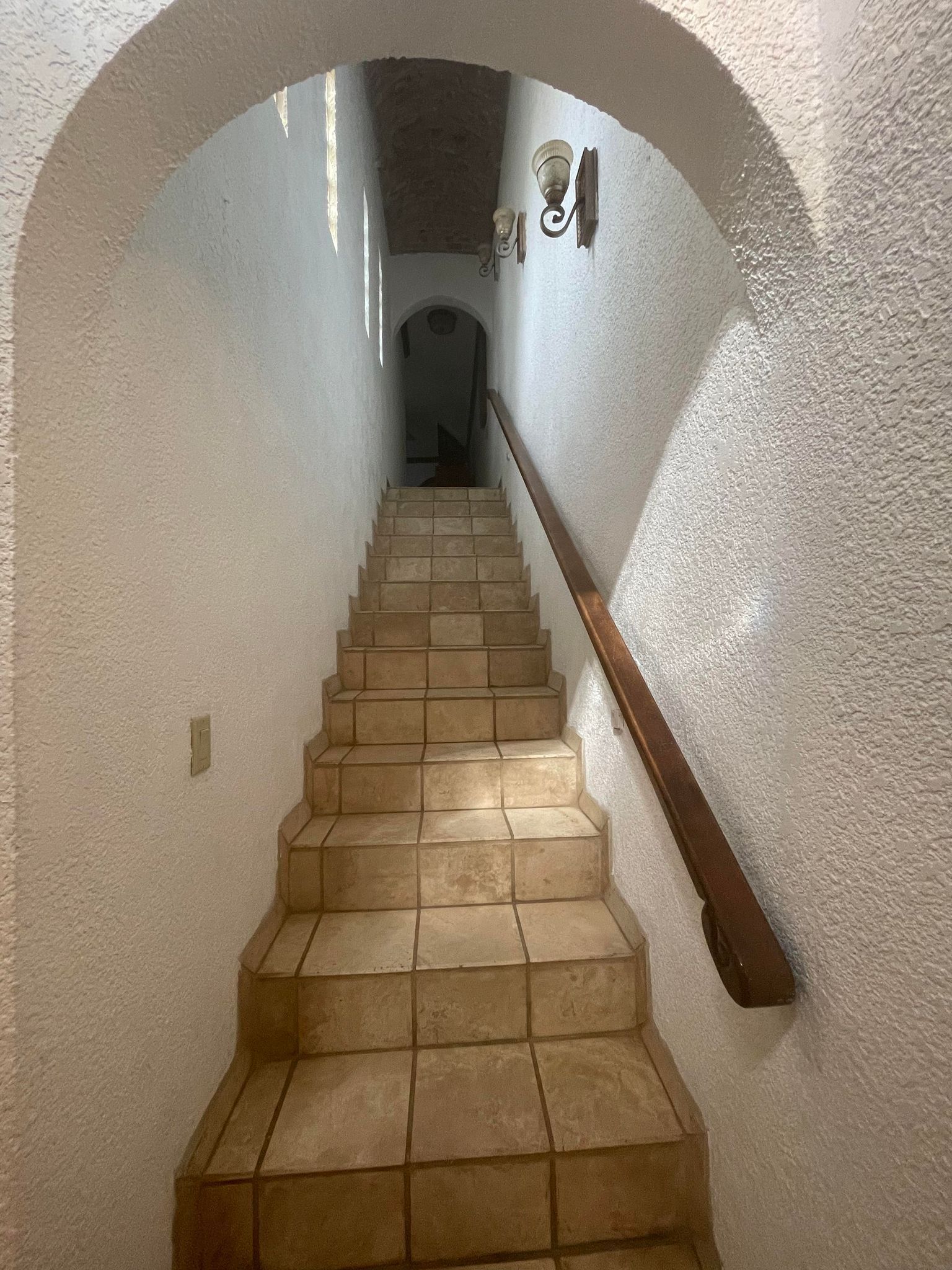 Un escalier recouvert de carreaux beiges, une main courante en bois et un mur blanc à la texture particulière.