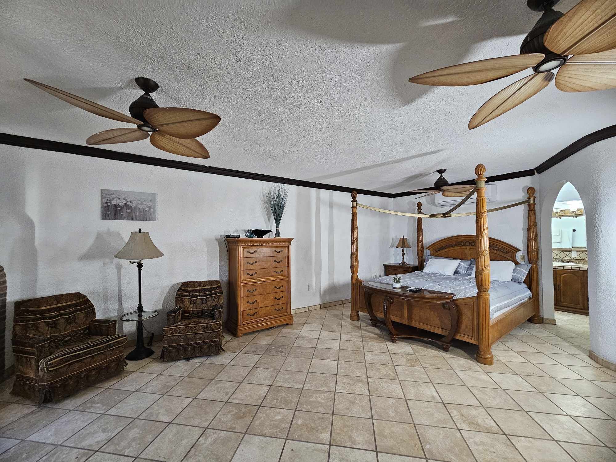 Un dormitorio amplio con una cama con dosel, un aparador de madera, una lámpara de pie y un ventilador de techo.