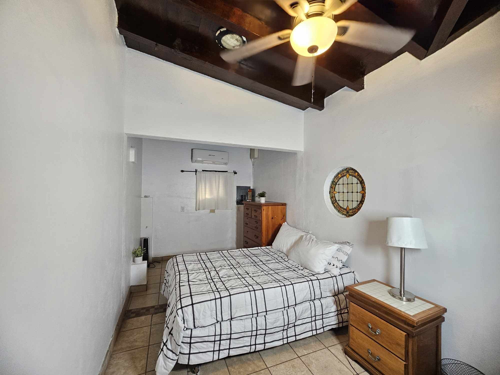 Un dormitorio con cama, cómoda de madera, ventilador de techo, aire acondicionado de pared y una ventana pequeña con cortina.