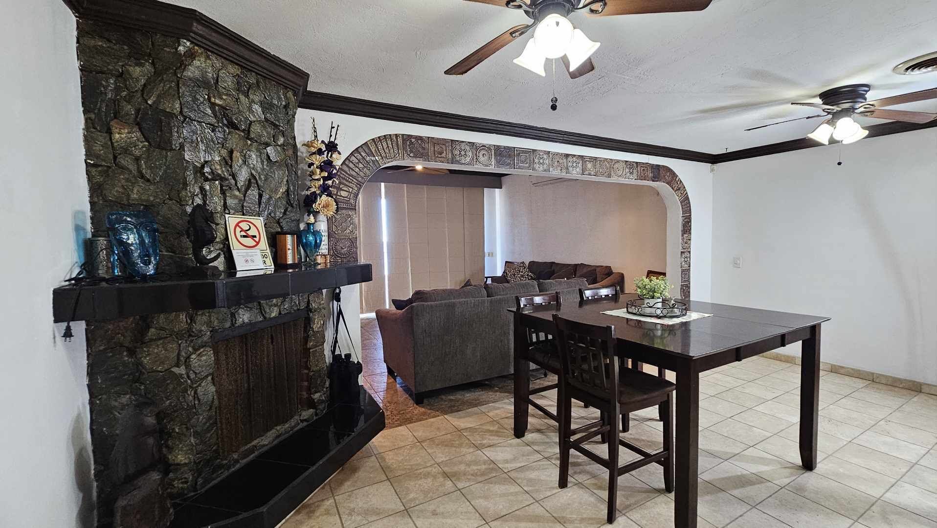 A living room with a stone fireplace, a dining table with chairs, a sofa, and a ceiling fan.