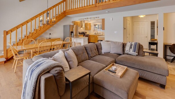 A modern living room with a large L-shaped sectional sofa, a wooden staircase, and a kitchen in the background. - Shuttle to Loon, Hot Tub & Pool! (Lincoln)