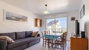 A living room with a sofa, dining table, chairs, a TV, and a balcony with a view. - Bright apartment with terrace in Erquy | 4 guests | Ref 531 (ERQUY)