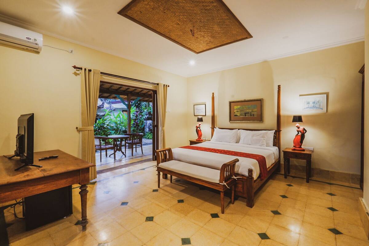 A hotel room with a bed, bedside tables, a desk with a television, and a view of a garden through an open door.