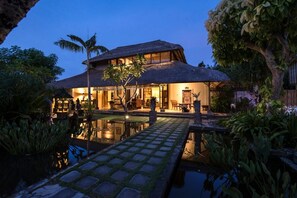 Una casa grande con techo de paja, piscina y abundante vegetación.