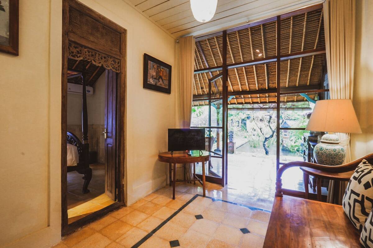 A room with a bed, a television, a small table, a chair, and a window with a view of greenery.