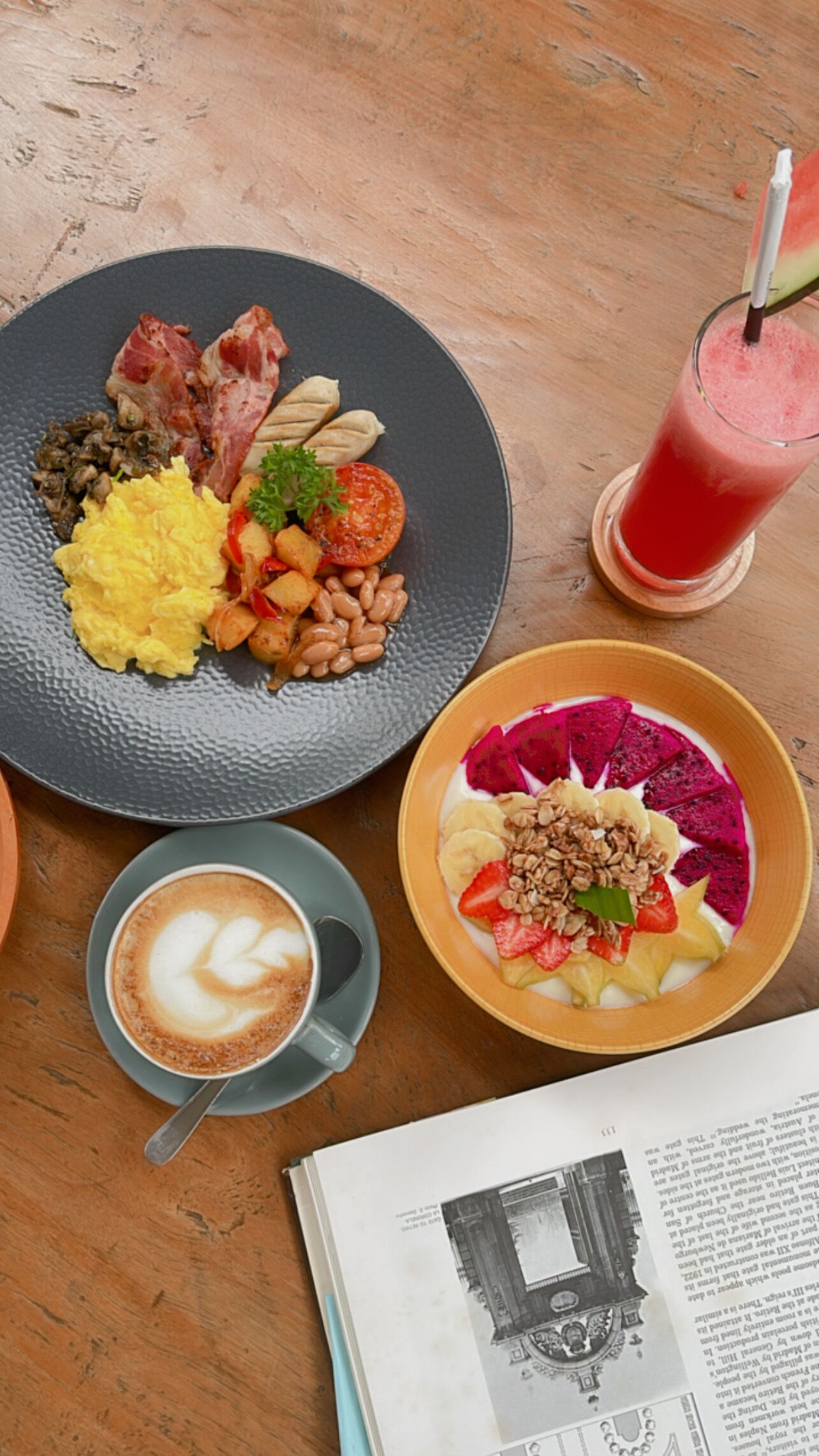 Una colazione con una piastra di uova strapazzate, pancetta, funghi e pomodori, una ciotola di yogurt con frutta e granola, un bicchiere di succo di frutta rossa e una tazza di caffè.