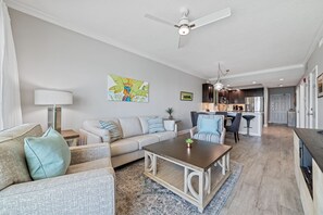 A modern living room with a sofa, coffee table, and a dining area with bar stools. There is a kitchen in the background with dark cabinets and stainless steel appliances. - Crystal Shores 1405 | Beach Front | Indoor Pool, Zero Entry Pool, Sauna and much more! (Gulf Shores)