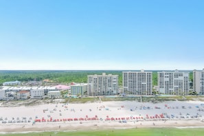 Vista de la playa con varios edificios altos, una playa arenosa con sombrillas y una zona verde.