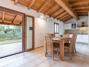 A dining area with a wooden table and chairs, a kitchen with white cabinets, and a view of trees through a sliding glass door. - Eucalipto by Interhome (Costa Rei)