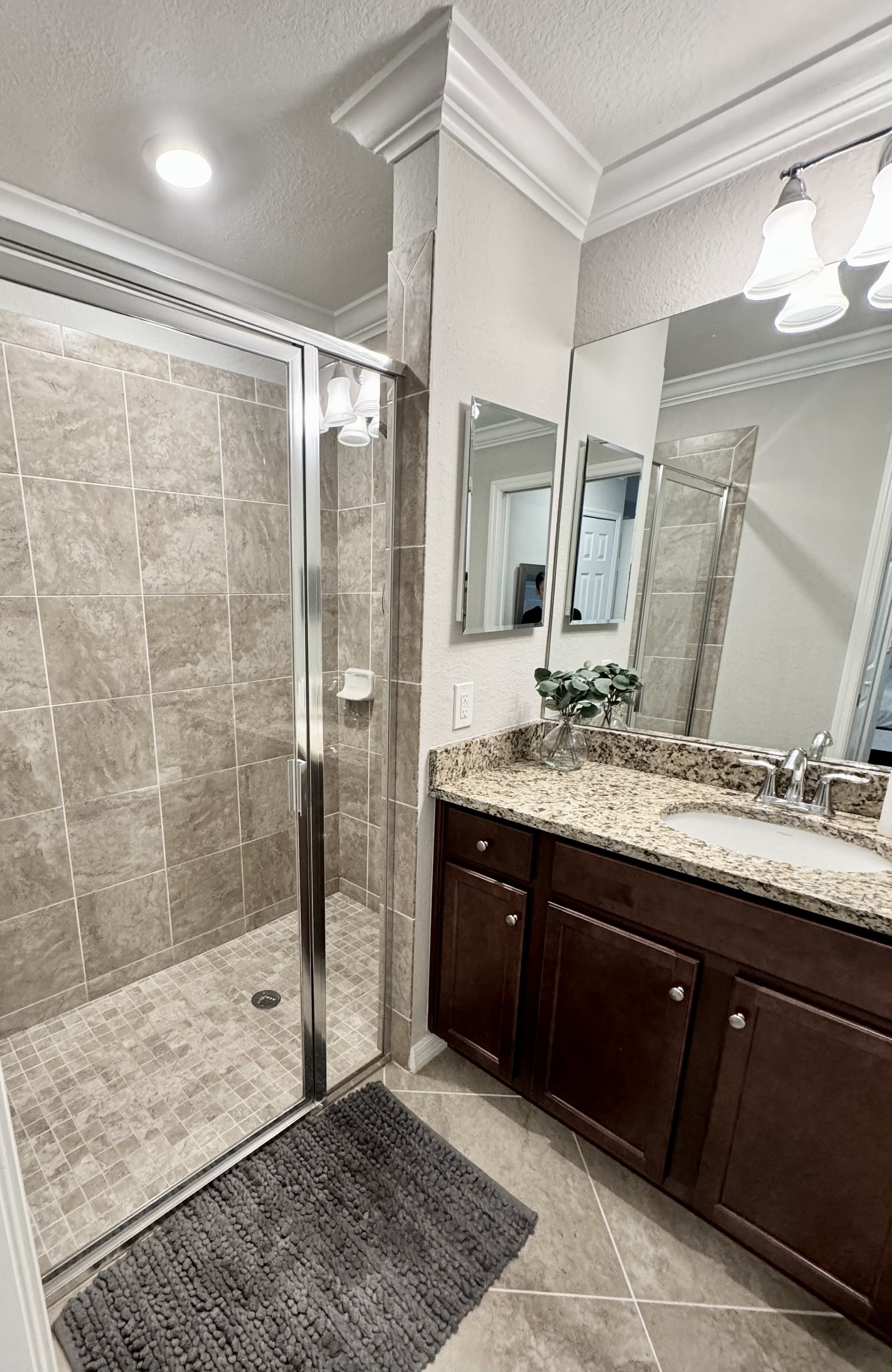 A bathroom with a glass-enclosed shower, a granite countertop sink, dark wooden cabinets, and a large mirror.