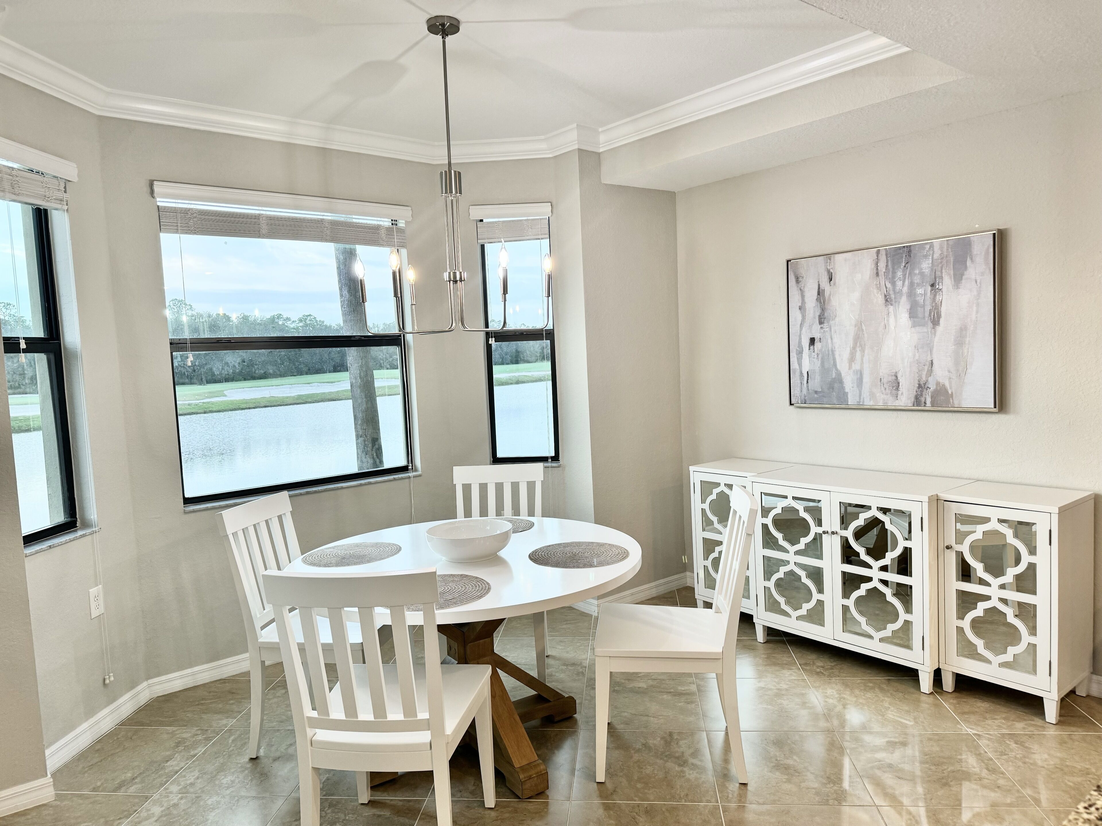 Un comedor con una mesa redonda, sillas blancas, un aparador blanco con puertas de cristal y un cuadro en la pared.