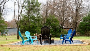 A rustic outdoor seating area with Adirondack chairs and a wooden table, surrounded by a natural landscape and a shed in the background.