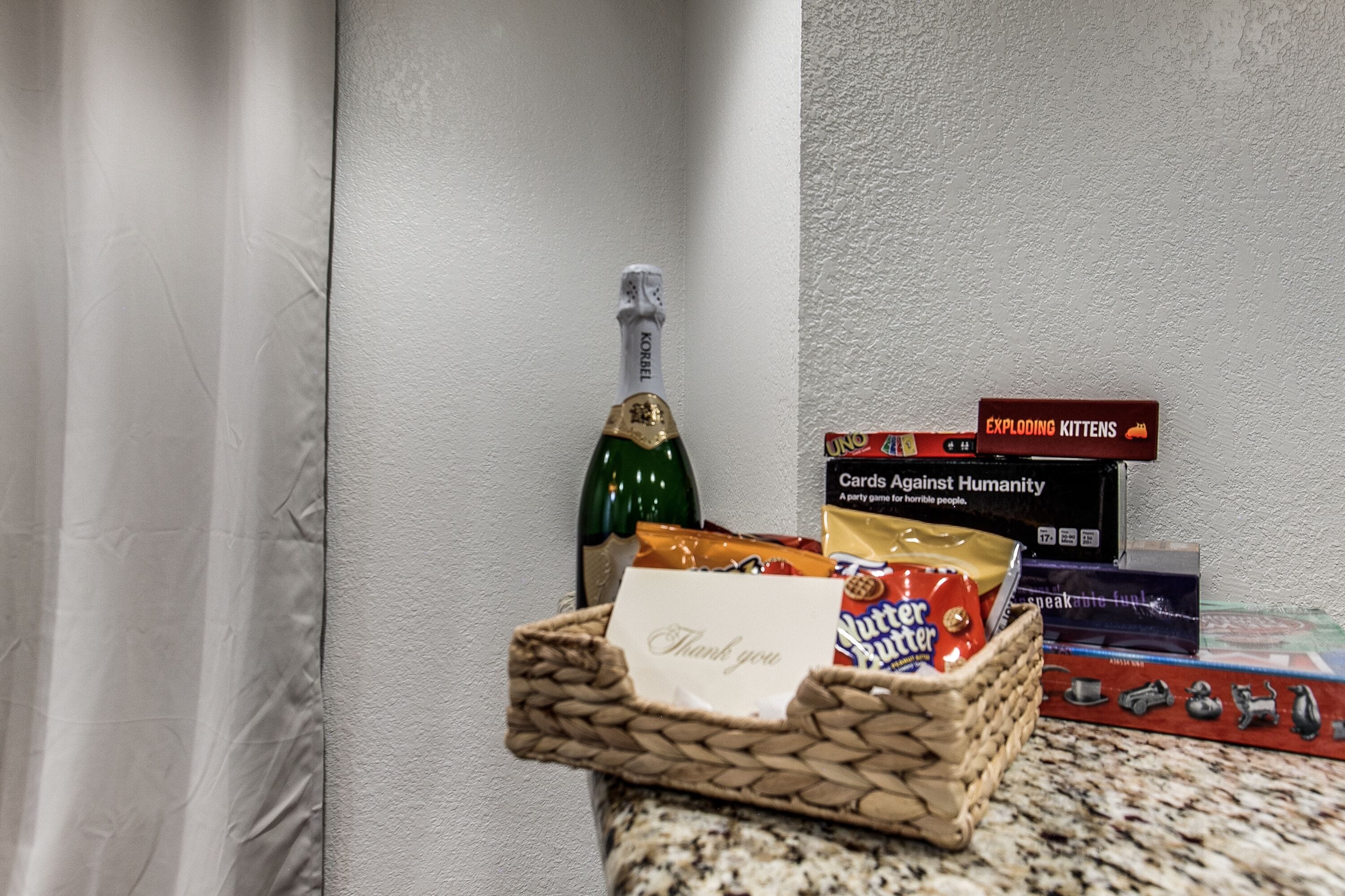 A basket with a thank you note, a bottle of champagne, and party games on a countertop.