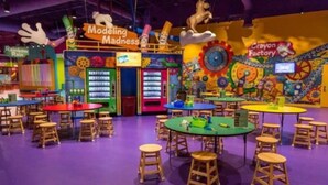 Area giochi al coperto dai colori vivaci, con tavoli e sgabelli, che include zone dedicate come 'Modeling Madness' e 'Crayon Factory'.