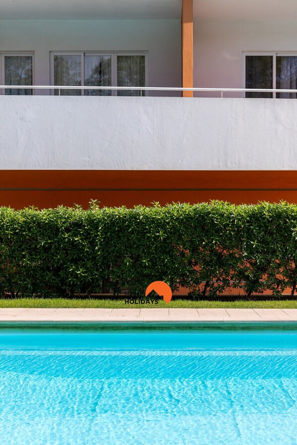 A swimming pool with a clear blue water surface, a hedge in the foreground, and a building with windows in the background. - #199 Flamingo 1108 by Home Holidays (Portimão)