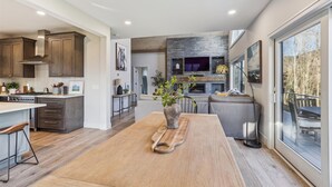 Una cocina moderna con una isla central, un comedor con una mesa grande y un espacio de sala de estar con chimenea y ventanas amplias.