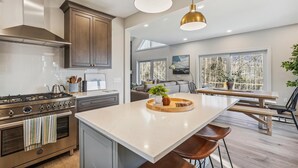 Una cocina moderna con una isla central, electrodomésticos de acero inoxidable y un comedor con vistas a árboles.