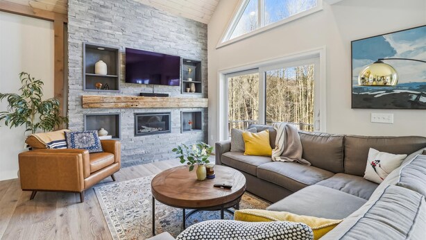 Un salón espacioso con pared de piedra, televisor grande, chimenea y vistas a árboles a través de amplias ventanas.