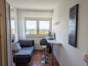 Un espacio habitable compacto con un sofá, una pequeña mesa con un portátil y vistas a una zona residencial a través de amplias ventanas.