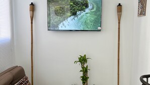 Ett vardagsrum med en brun soffa, en tv som är monterad på väggen, en liten växt i en trälåda och två höga golvlampor.