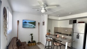 Ett modernt vardagsrum med en soffa, ett soffbord och en tv. Det finns också ett köksområde med ett kylskåp och ett matbord.