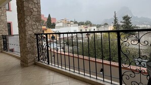 Un balcón con un pilar de piedra, suelo de baldosas y vistas a edificios y montañas a lo lejos.