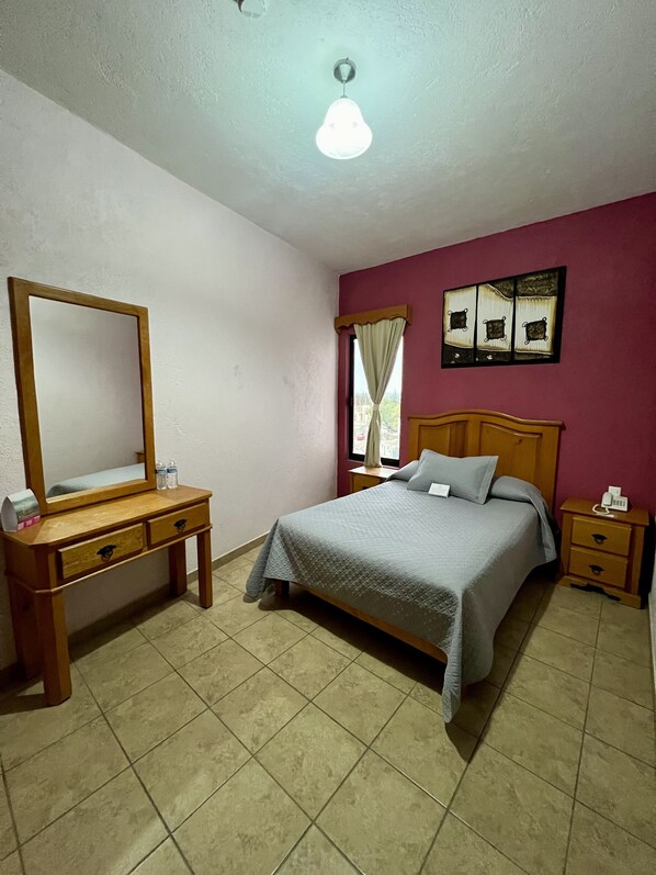 A bedroom with a bed, wooden nightstand, bedside table, mirror, and a wall with artwork.