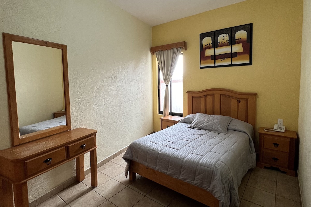 A bedroom with a wooden bed, a nightstand, a bedside table, a mirror, and a ceiling light.