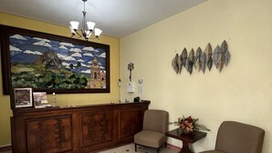 Un área de recepción con un escritorio de madera, dos sillas, una araña, un reloj de pared y un cuadro con un paisaje.