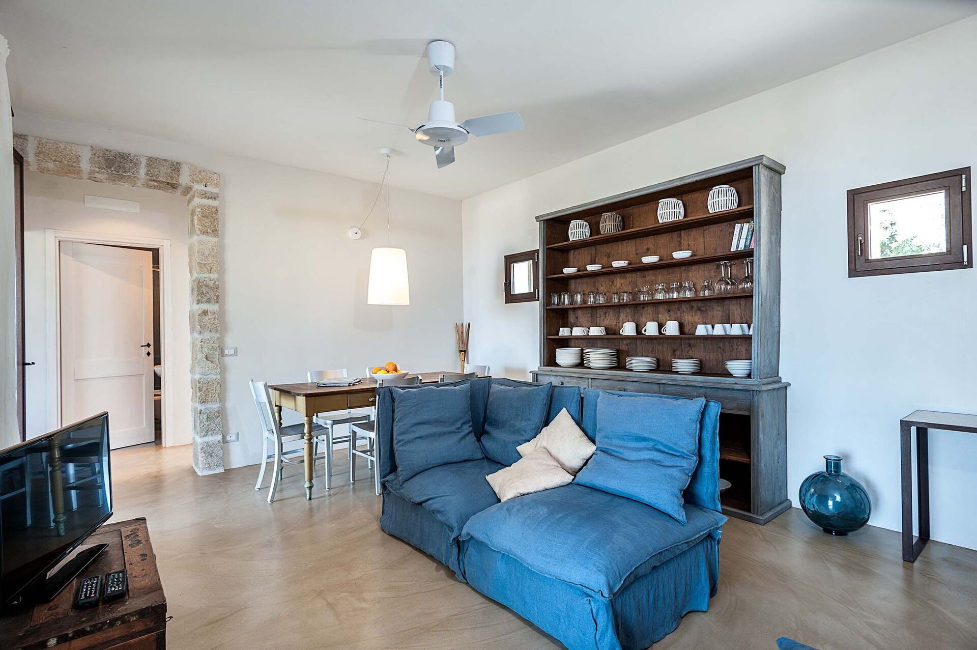 A living room with a blue sofa, a wooden coffee table, a dining area with a table and chairs, and a bookshelf filled with decorative items.