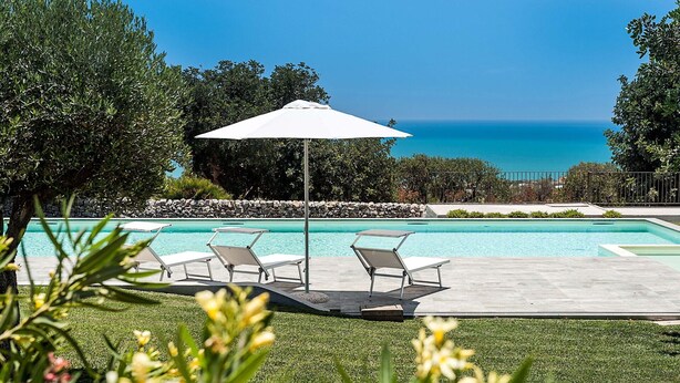 Ein Poolbereich mit Liegestühlen und Sonnensegel, umgeben von einer Steinmauer und Grünpflanzen, mit Blick auf einen klaren, blauen Himmel und das Meer.