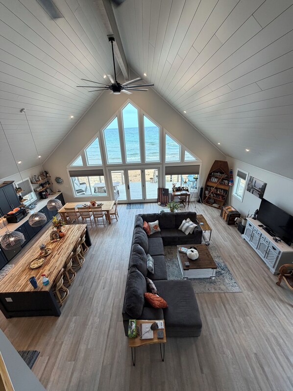 A spacious living area with a large window overlooking the beach, a dark-colored sofa, a wooden dining table, and a television.
