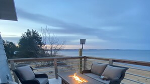 A deck with wicker furniture, a fire pit, and a view of the beach.