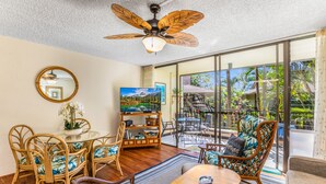 A spacious living area with a ceiling fan, a television, wicker furniture, and a large glass door leading to an outdoor patio. - 🌴AC | Garden View | 1st floor | Kalanikai #110🌴 (Kailua Kona)